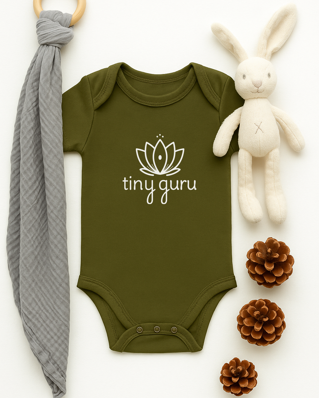Green baby onesie with 'tiny guru' logo, gray teething ring, white plush bunny, and pine cones on a white background