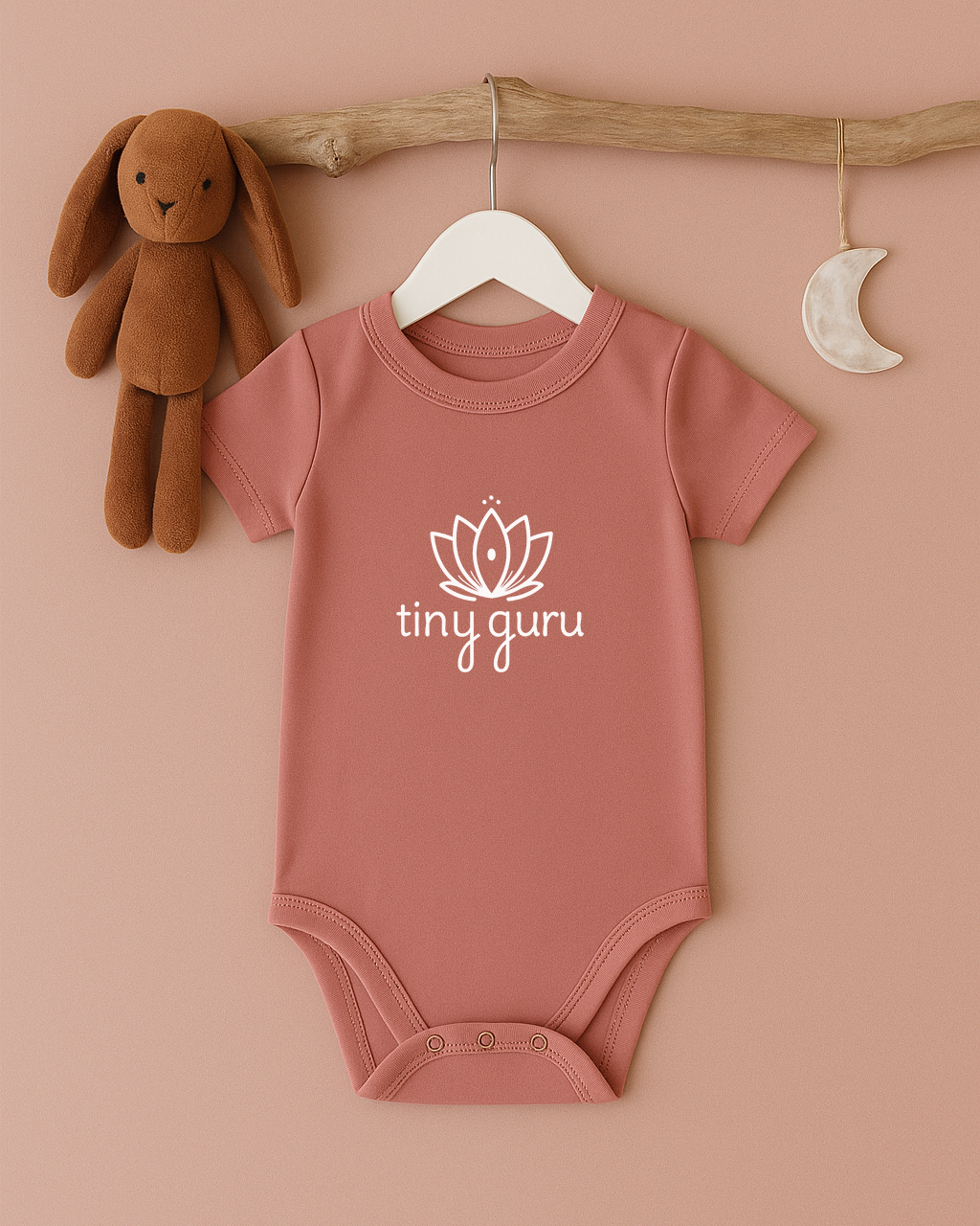 Pink baby onesie with 'tiny guru' logo on a hanger against a beige background