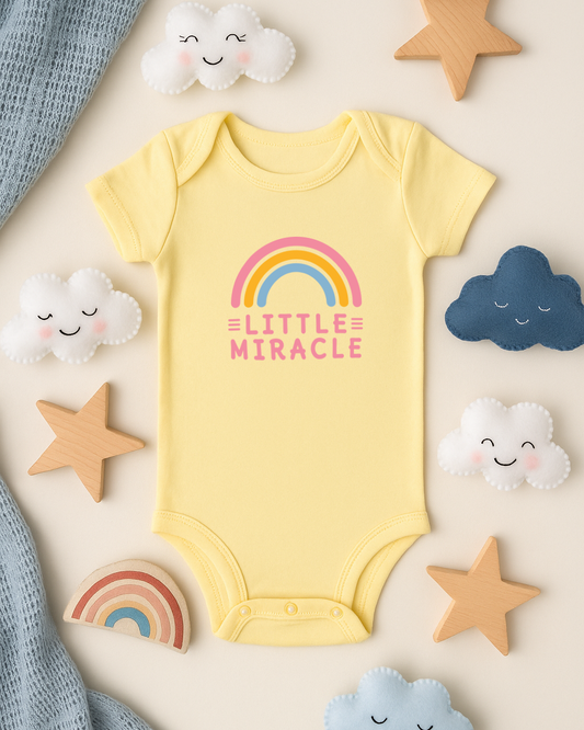 Yellow baby onesie with 'Little Miracle' print surrounded by decorative clouds and stars on a beige background
