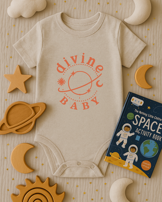 Beige baby onesie with 'divine BABY' print, surrounded by space-themed toys and a book on a beige background.
