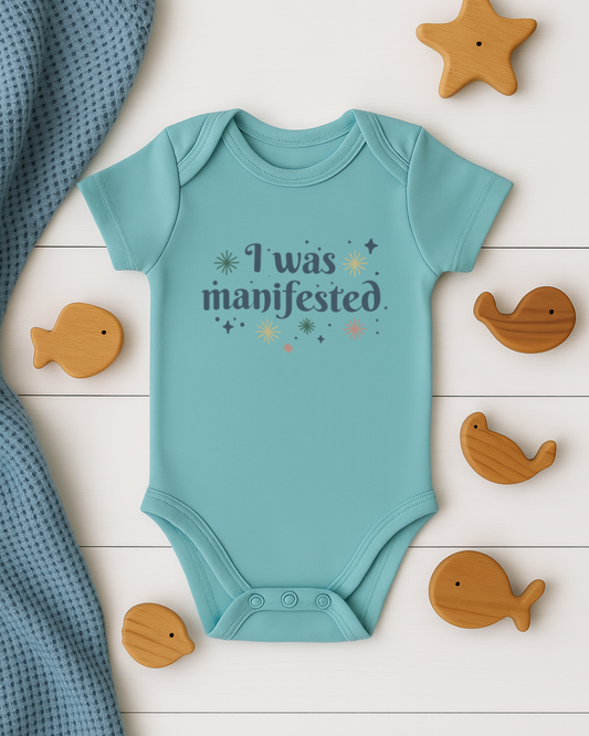 Teal baby onesie with 'I was manifested' text on a wooden surface with toys.