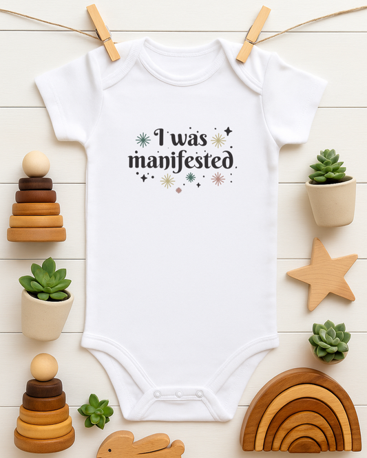 White baby onesie with 'I was manifested' text on a wooden surface with toys and plants.