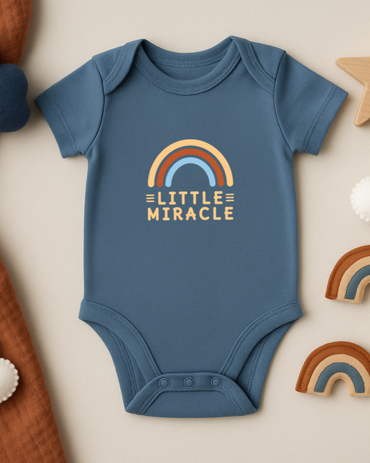 Blue baby onesie with 'Little Miracle' print on a beige background with decorative elements.