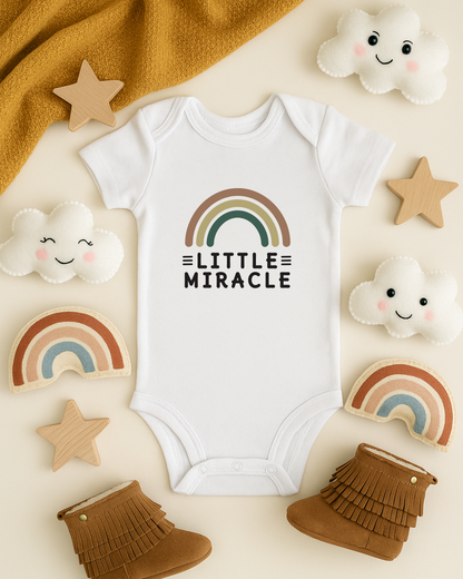 White baby onesie with rainbow design and 'Little Miracle' text, surrounded by cloud and star decorations on a beige background.
