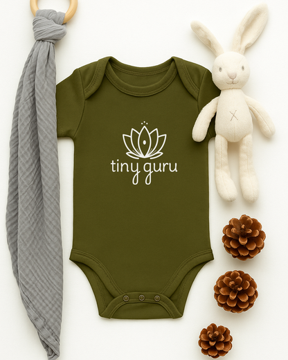 Green baby onesie with 'tiny guru' logo, gray teething ring, white plush bunny, and pine cones on a white background