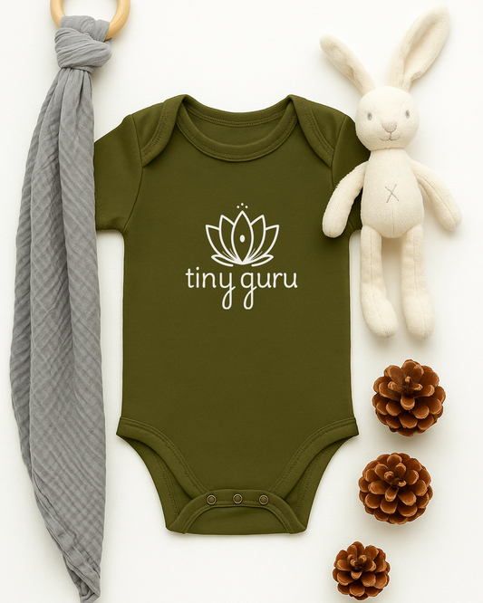 Green baby onesie with 'tiny guru' logo, gray teething ring, white plush bunny, and pine cones on a white background