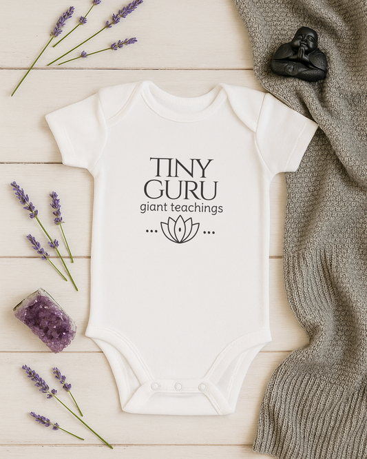 White baby onesie with 'Tiny Guru giant teachings' text on a wooden surface with lavender and a knitted blanket.