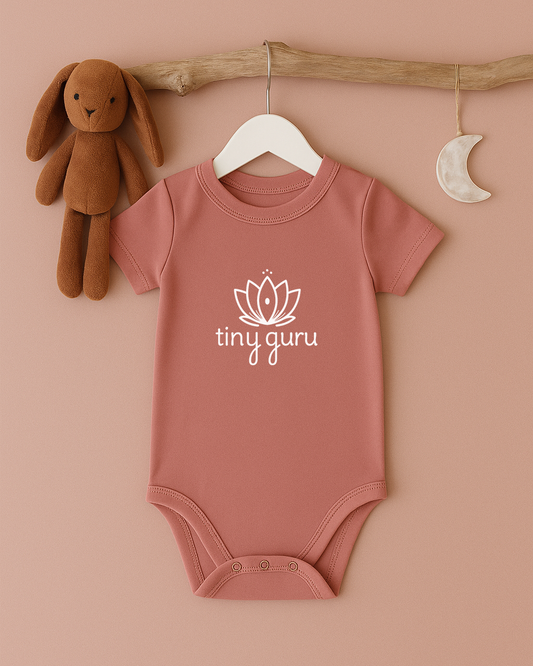 Pink baby onesie with 'tiny guru' logo on a hanger against a beige background